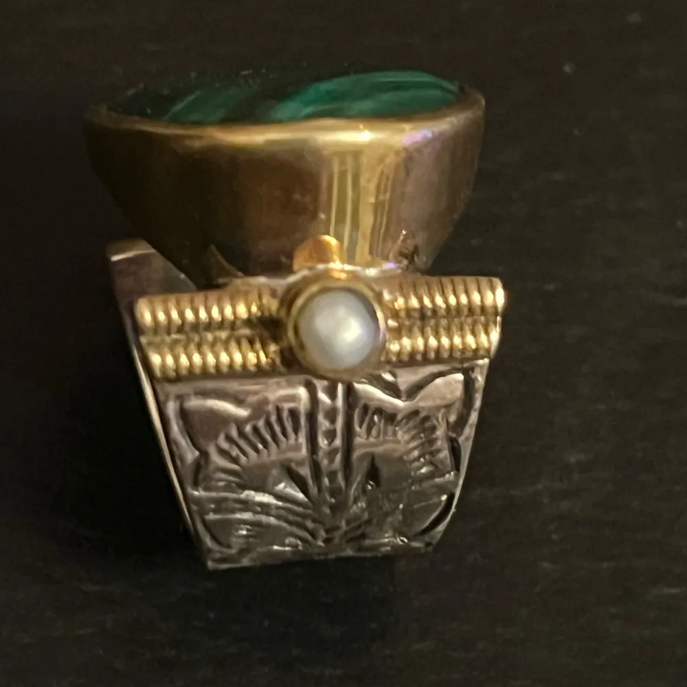 Unik ring från Canyon med en stor oval grön sten i centrum, omgiven av guldfärgad infattning. Sidorna pryds av små pärldetaljer och ett intrikat mönster på bandet. Perfekt för att ge en elegant touch till din stil.. Asusteet.