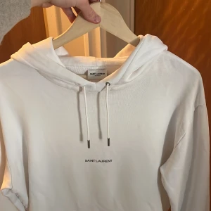 Saint Laurent hoodie  - Säljer en stilren vit hoodie från Saint Laurent med justerbar dragsko i huvan och en praktisk magficka. Perfekt för en avslappnad look.