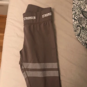 Bruna leggings från Stronger - Snygga bruna leggings från Stronger med hög midja och vita detaljer. Perfekta för träning med sitt stretchiga syntetmaterial och bekväma passform. Designade i Sverige med stilrena ränder och logotyp vid midjan.