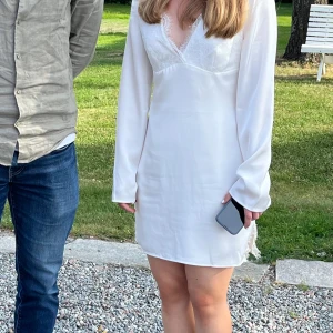 Vit silkesklänning  - Säljer en elegant vit klänning från NA-KD med spetsdetaljer och v-ringning. Klänningen har långa ärmar och en kort längd, perfekt för studenten och andra sommmartillställningar! Använd endast en gång och som ny i skick