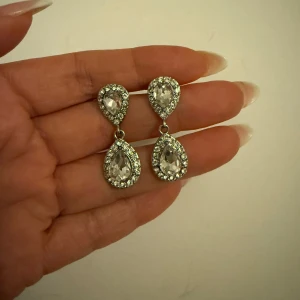 Glittriga örhängen med droppformade stenar - Eleganta örhängen i silverfärg med droppformade stenar omgivna av glittrande detaljer. Perfekta för att ge en lyxig touch till din outfit. De har en klassisk design som passar till många tillfällen.