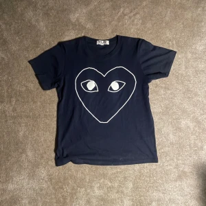 Mörkblå t-shirt från Comme des Garçons - mörkblå t-shirt från Comme des Garçons med ett unikt tryck av ett hjärta med ögon i vitt. T-shirten är kortärmad och gjord i mjuk bomull. Passar till nästan allt och kommer sitta perfekt till sommaren och våren.