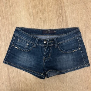 Mörkblå jeansshorts från Cherri Jeans - Snygga mörkblå jeansshorts från Cherri Jeans med dekorativa nitar på fickorna och en broderad detalj på bakfickan. Perfekta för en avslappnad stil. De har en knappgylf och klassisk femficksdesign.