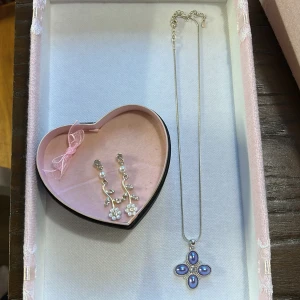 örhängen och halsband - Elegant smyckeset med ett par örhängen och ett halsband. Örhängena har blommotiv med pärlor och gnistrande stenar. Halsbandet har ett hänge med fyra lila stenar i en korsformad design. Perfekt för att ge en sofistikerad touch till din look. Vet ej vart nån av dem är ifrån eller om nån av dem är äkta då dem var min mammas gamla, så vi kan säga typ 100 kr för örhängena och 50 för halsbandet då dem ej kommer till användning 