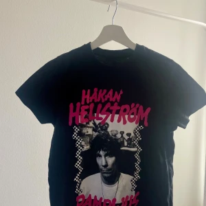 Svart t-shirt med Håkan Hellström-tryck - Svart t-shirt med Håkan Hellström-tryck i rosa och svartvitt. Perfekt för fans av artisten! T-shirten har korta ärmar och en klassisk rund halsringning. Använd enstaka gånger, nytt skick! 