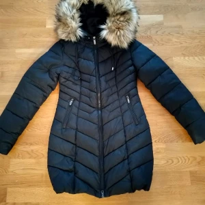 Svart dunjacka med pälsdetalj från Roco Baroco - Snygg svart dunjacka från Roco Baroco med en fluffig pälsdetalj på huvan. Jackan har en dragkedja framtill och två praktiska fickor med dragkedjor. Perfekt för kalla dagar!