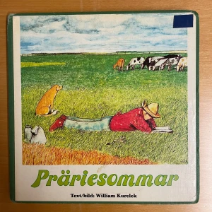 Präriesommar - Boken 'Präriesommar' av William Kurelek innehåller både text och illustrationer. Omslaget visar en person som ligger i gräset med en hund och kor i bakgrunden. Inuti boken finns illustrationer och berättelser om livet på en gård som på bilden en flicka i en ladugård med kor som ska mjölkas. 