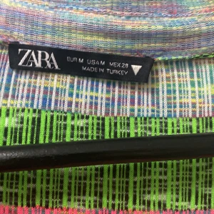 Färgglad randig klänning från Zara - Säljer en supercool och färgglad klänning från Zara i storlek M. Den har ett randigt mönster i grönt, rosa och grått med en snygg v-ringad design och knappar framtill. Klänningen är långärmad och har en åtsittande passform som framhäver figuren. Perfekt för en festlig kväll eller en dag när du vill sticka ut!