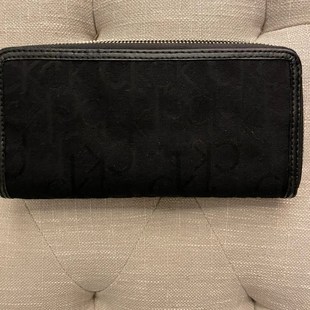 Säljer en snygg svart plånbok från Calvin Klein. Den har en stilren design med en silverfärgad metallplatta på framsidan där märket är ingraverat. Plånboken har en dragkedja och flera fack inuti för kort och kontanter. Nyskick. Laukut & Käsilaukut.