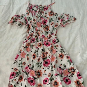Blommig sommarklänning från J. Stars - Säljer en superfin blommig klänning från J. Stars. Den har en offshoulder-design med korta ärmar och ett härligt blommönster i rosa och grönt på en vit bakgrund. 