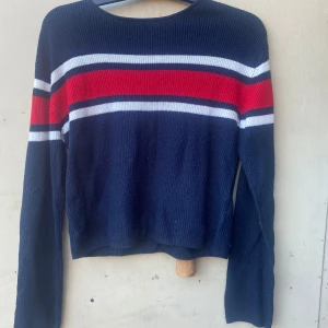 Mörkblå stickad tröja från H&M - Säljer en snygg mörkblå stickad tröja från H&M med ränder i rött och vitt över bröstet. Tröjan har långa ärmar och en normal passform, perfekt för både höst och vinter. Den är i bra skick och redo för nya äventyr!
