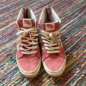 Rosa höga sneakers från Vans - Säljer ett par snygga rosa höga sneakers från Vans. De har en klassisk design med vita snören och den ikoniska vita sidoremsan. Skorna är i mocka och har en bekväm passform. Perfekta för en avslappnad stil!