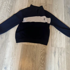 Svart och vit tröja med dragkedja som är sönder  - Snygg svart tröja med vit panel och dragkedja vid halsen. Perfekt för en stilren look. Tröjan har långa ärmar och en enkel design med en liten logotypdetalj på bröstet.dragkedjan är sönder 