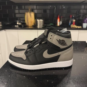  Air Jordans 1 high - Snygga Nike Air Jordans i svart och grått med klassisk design. Skorna har en hög siluett med snörning och en vit sula som kontrasterar mot det mörka lädret. Perfekta för en stilren look.