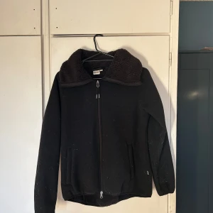 Svart teddyjacka från CRW - Mysig svart teddyjacka från CRW med dragkedja framtill. Jackan har långa ärmar och en hög krage för extra värme. Perfekt för kyliga dagar.