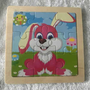 Trä Färgglatt kanin pussel - Charmigt pussel med en glad rosa kanin och en färgglad fjäril i bakgrunden. Pusslet har en träram och består av nio bitar som bildar en lekfull scen med blommor och en blå himmel.  Helt ny 