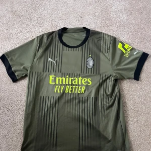 AC Milan tredje ställ.  - Säljer en olivgrön fotbollströja från Puma med korta ärmar och svarta detaljer. Tröjan har tryck med spelarnamnet 'R. Leão' med nummer 17 på ryggen. Tröjan är köpt från unisport och trycktes där.