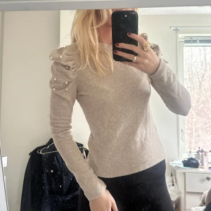 Beige tröja med pärldetaljer - Säljer en stilren beige tröja med långa ärmar och dekorativa pärlor på axlarna. Tröjan har en normal passform och är perfekt för en elegant look. 