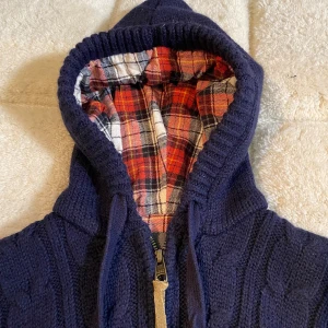 Cable knit wool cardigan - Säljer en snygg mörkblå stickad hoodie med dragkedja och rutigt foder i huvan. Tröjan är kabelskickad och ger då en italiensk design. Skjutit bra kvalitet. Storlek M på taggen men skulle säga sitter mer som S