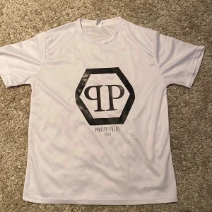 Vit t-shirt från Philipp Plein - Snygg vit t-shirt från Philipp Plein med ett stort svart logotryck på framsidan. Perfekt för en stilren och modern look. T-shirten har en rund hals och korta ärmar.                                                                                             Sitter större i storlek.                                                                Materialet är mer som en tränings tröja 