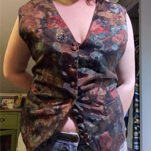 Blommig väst med knappar - Säljer en färgglad väst med blommigt mönster och knappar framtill. Västen har en v-ringad design. En av knapparna är lite trasig men det är inget som syns. Är xl men sitter som xxl