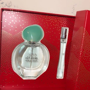 Acqua di Gioia parfymset från Giorgio Armani - Elegant parfymset med Acqua di Gioia från Giorgio Armani. Innehåller en flaska med en klar vätska och en mintgrön kork samt en mindre sprayflaska. Förpackningen är röd med gyllene detaljer och logotyp.