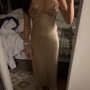 Champagnefärgad långklänning med volangdetaljer - Elegant champagnefärgad balklänning i satin med smala axelband och vackra volangdetaljer vid bysten. Klänningen har en figurnära passform och en stilren design som passar perfekt för bal! 