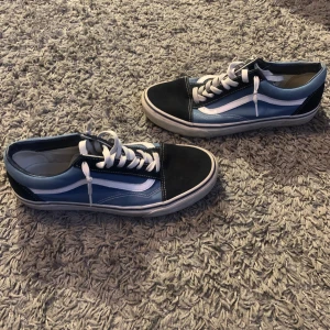 Blå och svarta sneakers från Vans - Säljer ett par snygga Vans sneakers i blått och svart med vita detaljer. Skorna har klassisk snörning och en vit sula med det ikoniska våffelmönstret. Perfekta för en avslappnad stil.