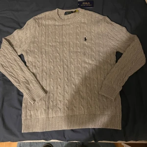 Ralph Lauren kabelstickad tröja - Säljer en vacker kabelstickad tröja från Polo Ralph Lauren. Tröjan har en elegant grå färg och rund halsringning samt ribbstickade muddar vid ärmslut och nederkant. På bröstet finns den ikoniska logotypen broderad i svart. Märke: Polo Ralph Lauren. Färg: Grå. Material: Högkvalitativ ullblandning. Storlek: Small. Skick: Mycket gott skick, helt ny. Perfekt för både vardag och fest. Pris kan diskuteras vid snabb och smidig affär. Kontakta mig för mer information.