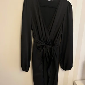 Svart klänning från Gina Tricot - Elegant svart klänning från Gina Tricot med långa ärmar och v-ringad design. Klänningen har en knytning i midjan som ger en smickrande passform. Perfekt för en stilren look.