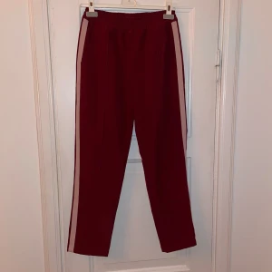 Vinröda kostymbyxor  - Snygga vinröda kostymbyxor med ljusrosa ränder längs sidorna. Perfekta för en avslappnad stil. Byxorna har två fickor och en elastisk midja för extra komfort.