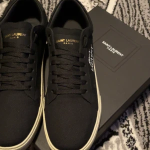 Svarta sneakers från Saint Laurent - Snygga svarta sneakers från Saint Laurent med vit sula och diskret logga på sidan. Skorna har klassisk snörning och är perfekta för en stilren look.Kvitto ingår och för frågor och funderingar så är det bara att ställa dom
