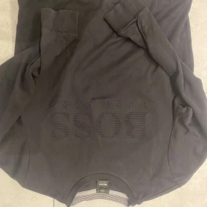 Svart tröja från Hugo Boss - Snygg svart tröja från Hugo Boss med broderad logga på framsidan. Tröjan har en slim fit passform och ribbade muddar vid ärmslut och nederkant. Oanvänd