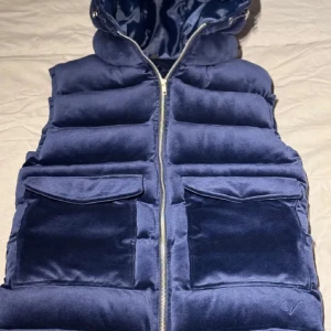 Mörkblå dunväst från Vendira - Snygg mörkblå dunväst från Vendiera med dragkedja och stora fickor framtill. Västen har en quiltad design och en huva för extra värme. Perfekt för kyliga dagar när du vill hålla stilen. Skick 10/10 använd 3 gånger skriv vid om ni undrar något! Pris kan diskuteras vid snabb affär!