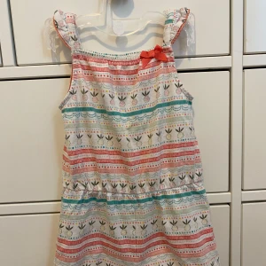 Färgglad sommarklänning med rosett - Söt och färgglad klänning med mönster i rött, turkos och vitt. Klänningen har breda axelband och en liten rosett på ena axelbandet. Perfekt för sommarens varma dagar!