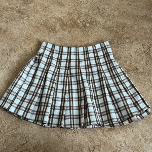 Rutig kjol från H&M Divided - Säljer en superfin rutig kjol från H&M Divided i brunt och vitt. Kjolen har en klassisk plisserad design som ger en snygg look. Perfekt för att skapa en stilren outfit!