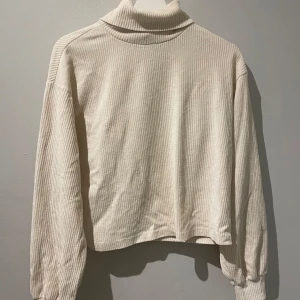 Beige ribbad polotröja - Säljer en beige polotröja med ribbad struktur. Tröjan har långa ärmar och en croppad passform. Den mjuka färgen gör den lätt att matcha med olika outfits, tyget är som manchestertyg.