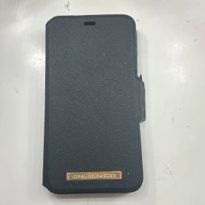 Svart mobilfodral från iDeal of Sweden - Snyggt svart mobilfodral från iDeal of Sweden med plats för kort. Fodralet har en stilren design med en magnetisk stängning och en guldig logotypdetalj på framsidan. Perfekt för att skydda din mobil med stil.