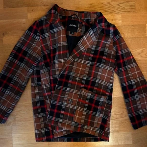 Rutig kavaj från Monki - Snygg rutig kavajjacka från Monki i brunt, svart och rött. Den har en klassisk krage och knäppning framtill med två knappar. Perfekta vårjackan!