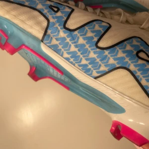 Vita fotbollsskor med blå detaljer från Nike - Snygga vita fotbollsskor från Nike med blå och rosa detaljer. Skorna har snörning och en modern design med en blå Nike-logga på sidan. Perfekta för fotbollsplanen med en stilren look. Använda en gång