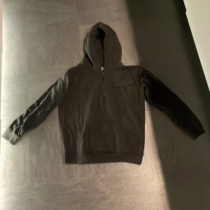 Svart hoodie från Lyle & Scott - Säljer en svart hoodie från Lyle & Scott Junior. Den har en praktisk dragkedja framtill och en känguruficka. På ärmen finns en broderad logotyp i gult. Perfekt för en stilren och bekväm look. Köpt på Lyle & Scott.