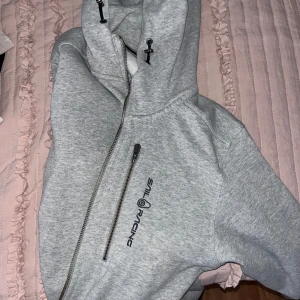 Grå hoodie från Sail Racing - Säljer en grå hoodie från Sail Racing med dragkedja och justerbar huva. Den har en bröstficka med dragkedja och logotyp på bröstet och ärmen. Perfekt för en avslappnad stil.