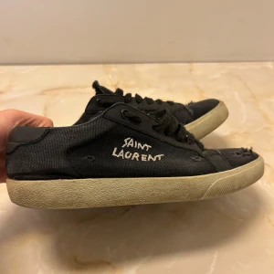 Svarta sneakers från Saint Laurent - Snygga svarta sneakers från Saint Laurent med en cool, sliten look. Skorna har en vit sula och svart snörning. Perfekta för en avslappnad stil med en touch av lyx. Vid frågor eller fler bilder, kontakta.
