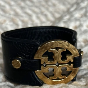 Svart Tor Burch läderarmband med gulddetalj  - Elegant svart, brett  läderarmband från Tory Burch. Armbandet har en stilren design med guldfärgade nitar på sidorna. Perfekt för att ge en lyxig touch till din outfit. Väl omhändertaget och i perfekt skick!