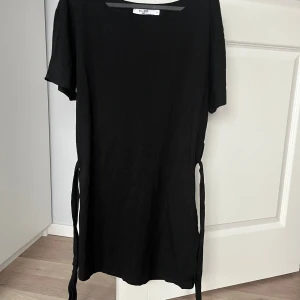 Svart klänning från NA-KD - Snygg svart klänning med korta ärmar och knytband i midjan. Klänningen har en enkel design som gör den lätt att matcha med olika accessoarer.