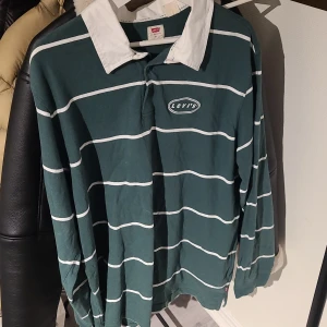 Randig pikétröja från Levi's - Säljer en snygg grön och vit randig pikétröja från Levi's. Tröjan har en klassisk krage i vitt och långa ärmar. Perfekt för en avslappnad stil. Levi's-loggan är broderad på bröstet.