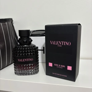 Valentino Uomo Born in Roma Intense - Säljer en elegant parfymflaska från Valentino, Uomo Born in Roma Intense. Flaskan har en lyxig design med svart och rosa detaljer och en texturerad yta. Perfekt för den som vill ha en intensiv och sofistikerad doftupplevelse.