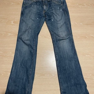 Bootcut / Flare Guess Jeans - Säljer ett par klassiska blå jeans från Guess med bootcut-stil. De har en snygg vintage-look med lätt slitna detaljer och en ikonisk Guess-logga på bakfickan. Perfekta för en avslappnad stil.