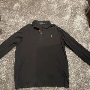 Svart pikétröja från Ralph Lauren - Säljer en klassisk svart pikétröja från Ralph Lauren. Tröjan har långa ärmar och en stilren krage med knappar. Perfekt för en avslappnad men ändå stilfull look.