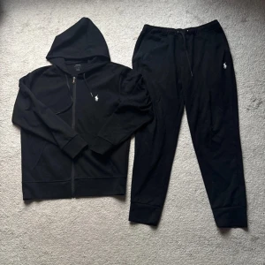 Svart hoodie och byxor från Ralph Lauren - Säljer en äkta stilren svart hoodie med dragkedja och matchande byxor från Ralph Lauren. Hoodien och byxorna qr kod och en liten broderad logga på bröstet och byxorna har en dragsko i midjan för justerbar passform. Perfekt för en avslappnad look.